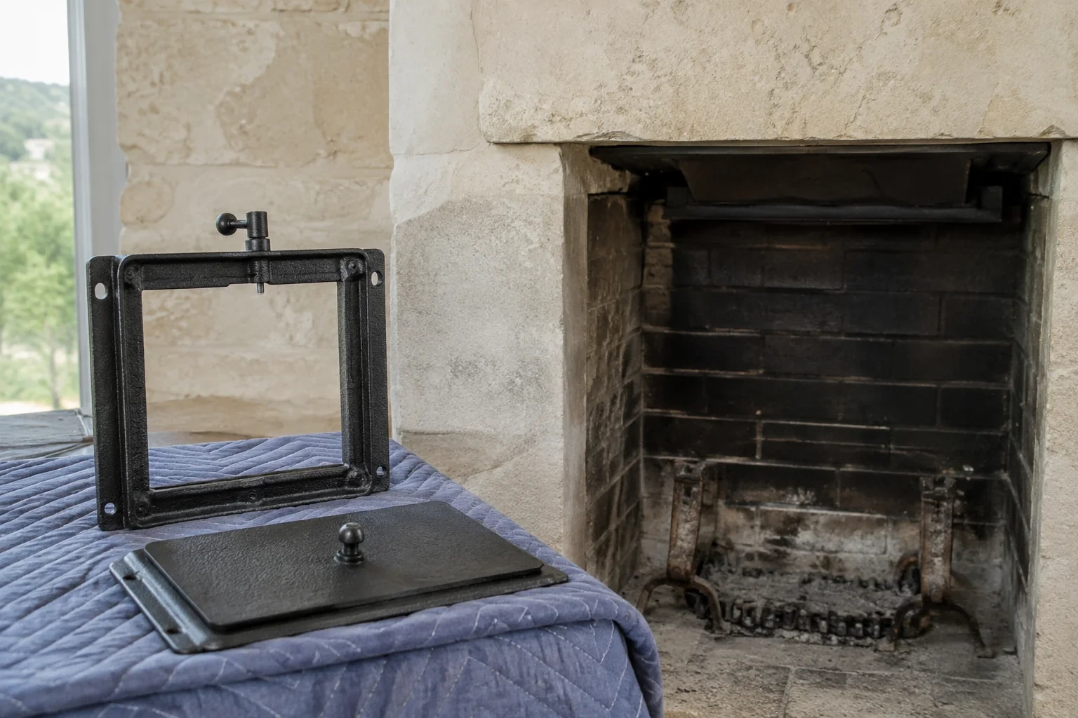 Black rectangular metal frame and matching panel resting on a padded cloth beside an open brick-lined fireplace.